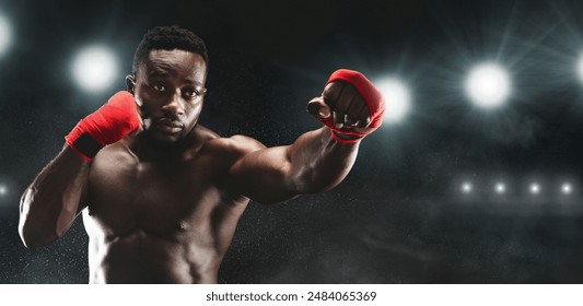 A muscular boxer, wearing red hand wraps, throws a punch in a boxing ring. The lights shine brightly above him, creating a dramatic atmosphere. - Powered by Shutterstock