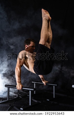 Muscular build man doing calisthenics on parallels bar indoor on black, smoked background. concept of healthy lifestyle and power