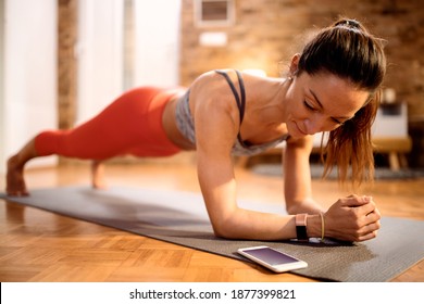 Muscular Build Woman Exercising Strength In Plank Pose And Looking At Timer On Her Smart Phone At Home. 