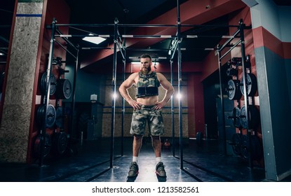 Muscular Caucasian Bearded Man Standing In Military Style Weighted Vest In Gym. Weight Plates, Kettlebells, Barbell And Crossfit Tires In Background.