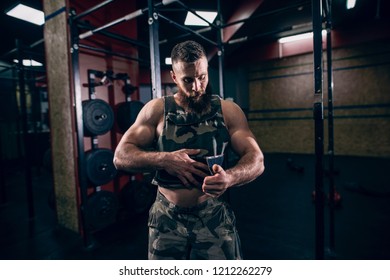 Muscular Caucasian Bearded Man Tightening Up Military Style Weighted Vest In Gym. Weight Plates And Kettlebells In Background.