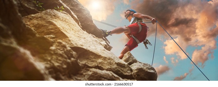 Muscular climber man in protective helmet abseiling from cliff rock wall using rope Belay device and climbing harness on evening sunset sky background. Active extreme sports time spending concept. - Shutterstock ID 2166630517
