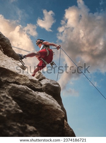 Muscular climber man in protective helmet abseiling from cliff rock wall using rope Belay device and climbing harness on evening sunset sky background. Active extreme sports time spending concept.