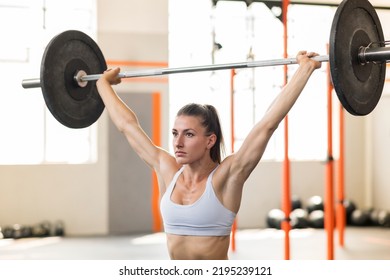 Muscular Female Athlete With Ponytail Looking Away And Doing Snatch Exercise With Heavy Barbell During Intense Weightlifting Workout On Blurred Background Of Sunlit Gym