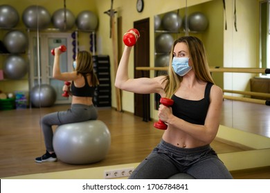 Muscular Female Fitness Sports Trainer Coach In White Medical Mask Show Biceps With Red Dumbbell Invite To Do Online Fitness Class Sitting On Yoga Ball In Empty Gym. Athletes Strong To Stop COVID-19