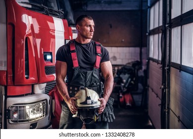 Muscular fireman holding a protective helmet in a garage of a fire department, standing next to a fire engine and looking outside