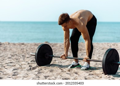 Muscular guy bodybuilder doing exercises with big dumbbells outdoors on the beach, on a summer sunny day, training on the sea coast on the background