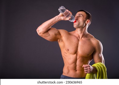 Muscular Guy - Bodybuilder Posing On A Gray Background. Athletic Man Holding A Bottle Of Water In Hand, A Towel Around His Neck. Sport, Health, Bodybuilding, Strength, Power - A Concept Sports.