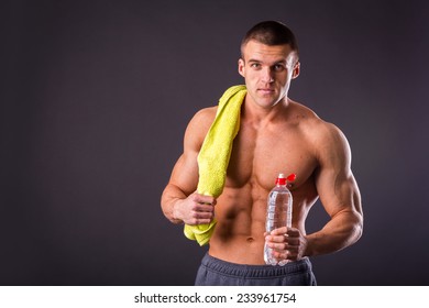 Muscular Guy - Bodybuilder Posing On A Gray Background. Athletic Man Holding A Bottle Of Water In Hand, A Towel Around His Neck. Sport, Health, Bodybuilding, Strength, Power - A Concept Sports.