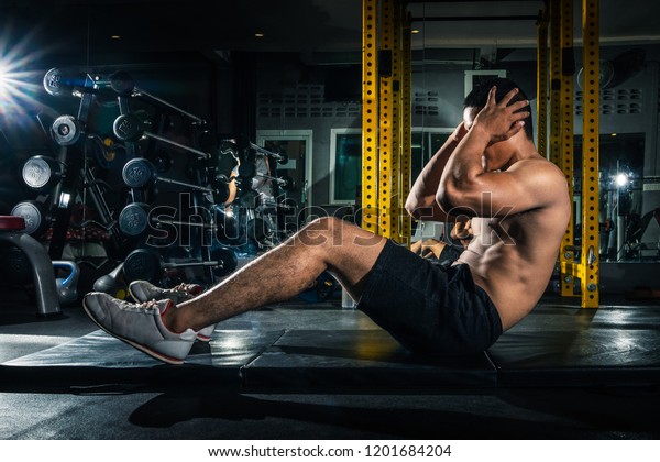 Muscular guy doing sit ups at gym with\
other people in background. Young athlete doing stomach workout in\
modern gym. Handsome fit man doing crunches at\
gym.