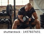 Muscular guy lifting dumbbell while sitting on bench at gym. Mature african athlete using dumbbell during workout. Strong man under physical exertion pumping up bicep muscle with heavy weight.