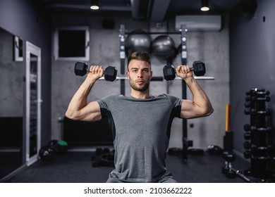 Muscular guy lifting dumbbell while sitting on a bench at the gym. A young athlete using dumbbells during a workout. Strong man under physical exertion pumping up bicep muscle with weight - Shutterstock ID 2103662222
