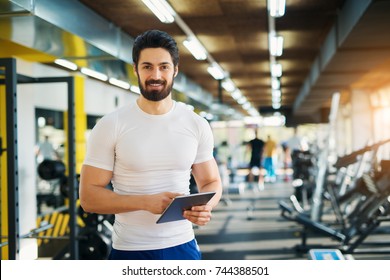 Muscular Handsome Bearded Trainer Looking At The Camera While Standing With A Tablet In The Gym.