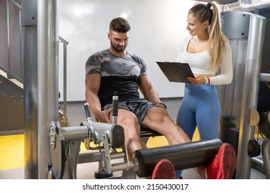 Muscular Handsome Man Doing Exercises On Leg Press Machine And Female Personal Trainer With Clipboard Calculating His Muscle Progress During Workout Class At Gym.