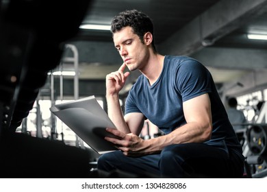 Muscular Handsome Trainer Looking At Fitness Plan On Clipboard For Working Out In The Fitness Gym.