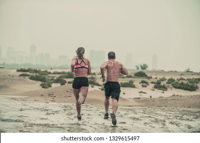 Muscular Male And Female Athlete Covered In Mud Running Down A Rough Terrain With A Desert Background In An Extreme Sport Race With Grungy Textured Finish