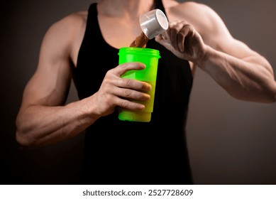 A muscular man in a black tank top pours chocolate protein powder into a green shaker. Shot against a dark background, the image represents fitness, sports nutrition, and bodybuilding themes. - Powered by Shutterstock