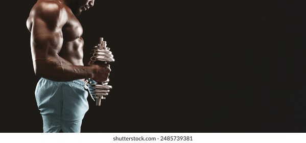 A muscular man in blue shorts lifts a heavy dumbbell while standing in a dark room. - Powered by Shutterstock