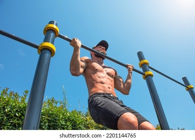 Muscular Man In A Cap Doing Reverse Pull-up On The Horizontal Bar In The Park Outdoors