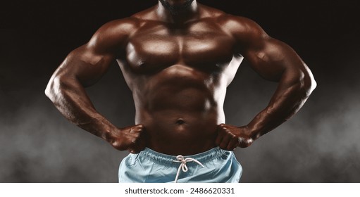 A muscular man with defined biceps, triceps, and chest poses with hands on his hips against a dark, smoky background. - Powered by Shutterstock