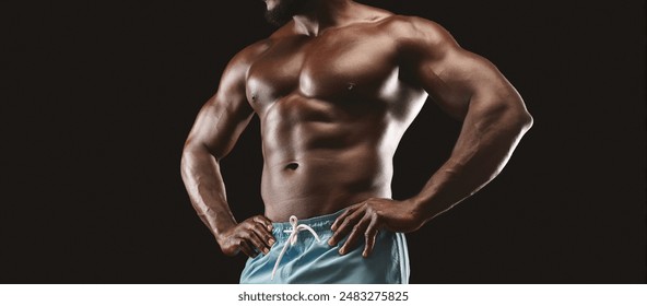 A muscular man with a defined physique poses with his hands on his hips against a black background. - Powered by Shutterstock