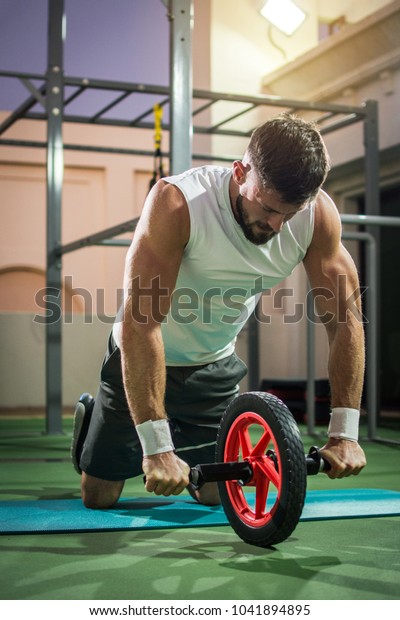 Muscular man doing exercises with abdominal toning\
wheel at gym.