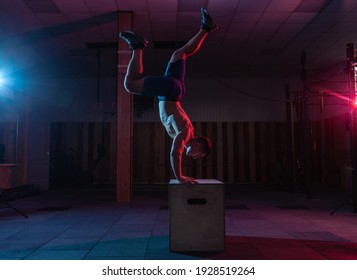 Muscular Man Exercises A Handstand On A Wooden Box In A Dark Gym. Athlete Workout In Red Blue Neon Light. Cross, Functional Training