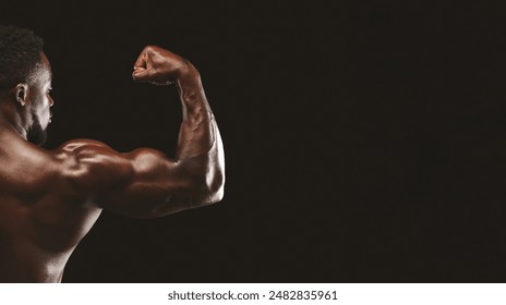 A muscular man flexes his bicep against a black background, showcasing his strength and fitness. - Powered by Shutterstock