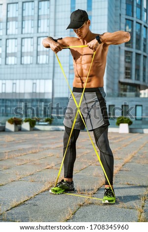 Muscular man performs exercises using a resistance band. Photo of young man training in the city. Strength and motivation. Full length