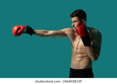 A muscular man is practicing boxing by throwing punches indoors against a solid blue background. He is wearing red boxing gloves and is shirtless, showcasing his toned physique and focused expression. - Powered by Shutterstock