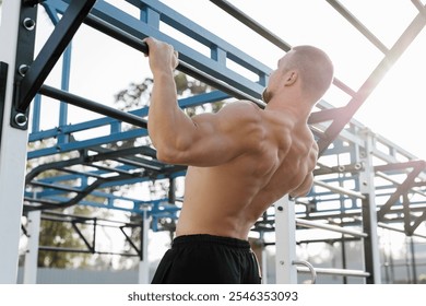 A muscular man pulls himself up on a horizontal bar in an outdoor park at sunset