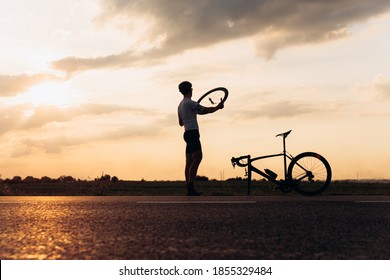 Muscular Man In Silhouette Fixing Wheel On Broken Bike On Fresh. Natural Landscape And Amazing Sunset. Sportsman Training Outdoors.