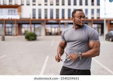 Muscular man standing outdoors in a shopping area. A muscular man holds sunglasses while standing in a shopping area, showing confidence and strength during the day. - Powered by Shutterstock