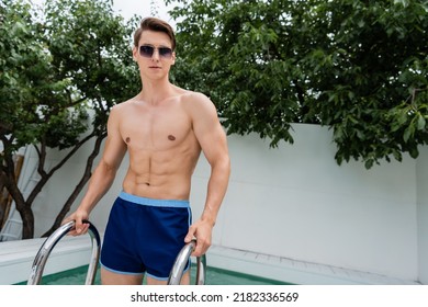 Muscular Man In Sunglasses And Swimming Trunks Standing Near Pool Ladder