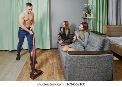 A Muscular Man Is Tidying Up The Room With Vacuum Cleaner. Two Young Women Watch As An Athletic Male Cleans The Floor In Apartment. Striptease Performer  Impersonates A Cleaner At A Hen Party In Dorm.