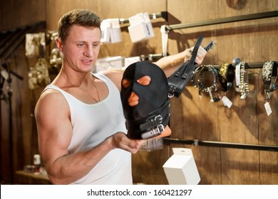 A Muscular Man In A Vest Holding A Leather Whip And Head In Mask In The Wooden Room