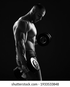 Muscular Men Bodybuilder Is Working Out In Gym, Lifting Alternately Two Dumbbells, Doing Exercises For Biceps And Looking Down Over Black Background. Young Man Lifting Weights. Black And White 