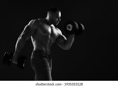 Muscular Men Is Working Out In Gym, Lifting Alternately Two Big Heavy Dumbbells, Doing Exercises For Biceps Over Black Background With Copy Space. Young Man Lifting Weights. Black And White 