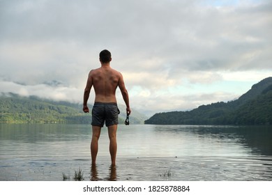 A Muscular Mid 20s Male Stands Confidently After Wild Swimming In Loch Lomond, Scotland. Wearing Swimming Trunks And With Swimming Goggles In Hand. Picturesque Views All Around. Relaxing.