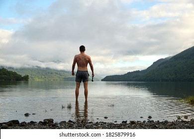 A Muscular Mid 20s Male Stands Confidently After Wild Swimming In Loch Lomond, Scotland. Wearing Swimming Trunks And With Swimming Goggles In Hand. Picturesque Views All Around. Relaxing Wide Shot.