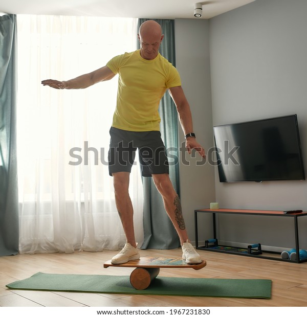 Muscular middle aged\
caucasian man standing on balance board installed on room floor.\
Fitness at home\
concept