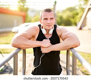 Muscular Millennial Man Having A Break Between The Workout Exercises And Leaning On A Dip Bar In Outdoor Public Fitness Park  With Headphones On His Neck - Active And Healthy Lifestyle Concept