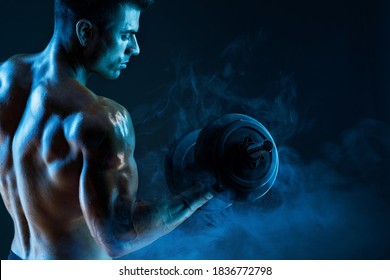 Muscular model sports young man with dumbbells in hand on dark background. Fashion portrait of strong brutal guy. Sexy torso. Male flexing his muscles.