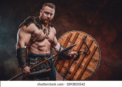 Muscular and nude viking with fur and beard holding spear and shield in dark red background.