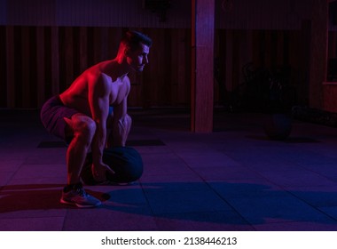 A Muscular Powerful Man With A Naked Torso Exercises With A Heavy Bag In A Red Blue Neon Light. Cross Workout In A Modern Gym