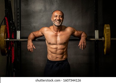 Muscular shirtless mature older bodybuilding athlete with balding gray hair  leaning against a barbell in a gym smiling at the camera