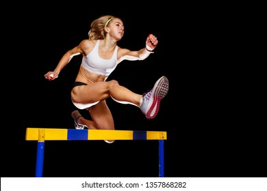 Muscular sportswoman jumping over hurdle on sprint race isolated on black background. Track and field event concept image - Powered by Shutterstock