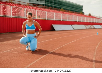 Muscular sporty woman in a blue sportswear and sneakers kneeling on one leg on the running track at the stadium outdoors and preparing for a race or training. Healthy lifestyle concept. Copy space. - Powered by Shutterstock