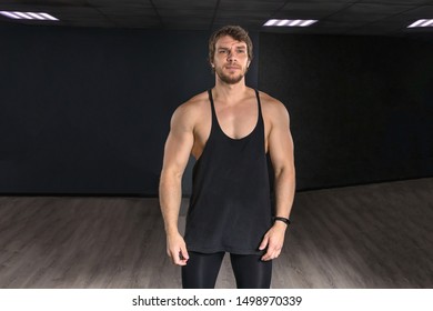 Muscular Strong Athletic Guy In A Black Tank Top Standing Posing Near Dark Wall. Concept Sport Photo With Copy Space