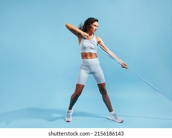 Muscular Woman Exercising With Elastic Band. Female In Fitnesswear Pulling Resistance Band On Blue Background.